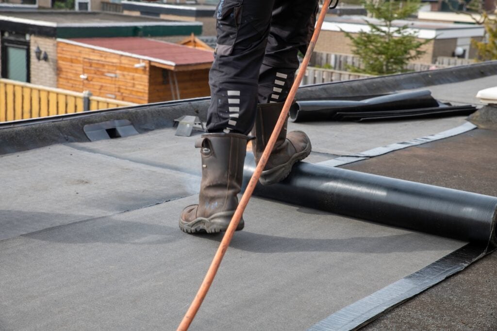 Roofer standing on a flat residential roof using a propane torch to heat and install roll roofing membrane for waterproofing and roof repair. Scene shows work boots, tools and neighborhood houses.