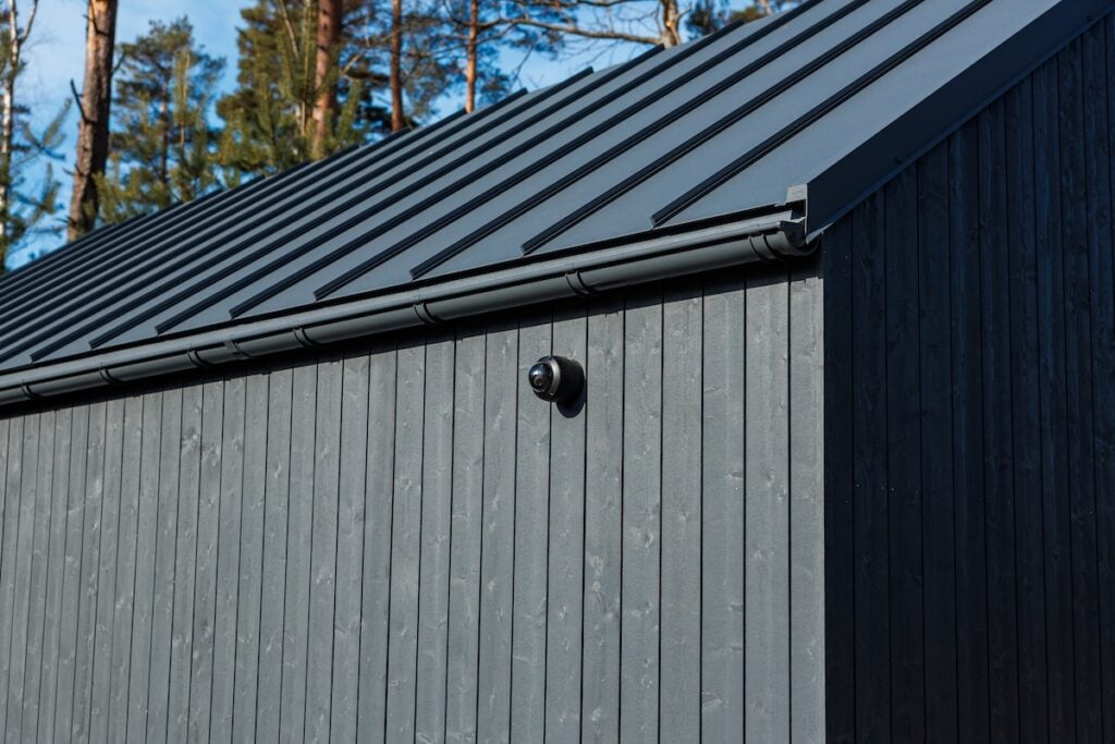 Charred timber wall with vertical cladding supports a matte metal roof and gutter. A dome camera sits on the eave. Conifer trunks and daylight suggest a wooded setting.
