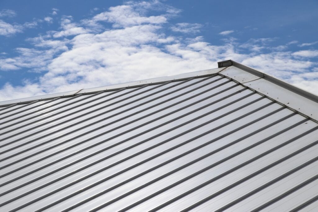 Close-up view of a silver metal roof with vertical ridges against a blue sky with scattered white clouds, highlighting the long-term value and distinctive appearance that are among the key pros and cons of choosing a metal roof.