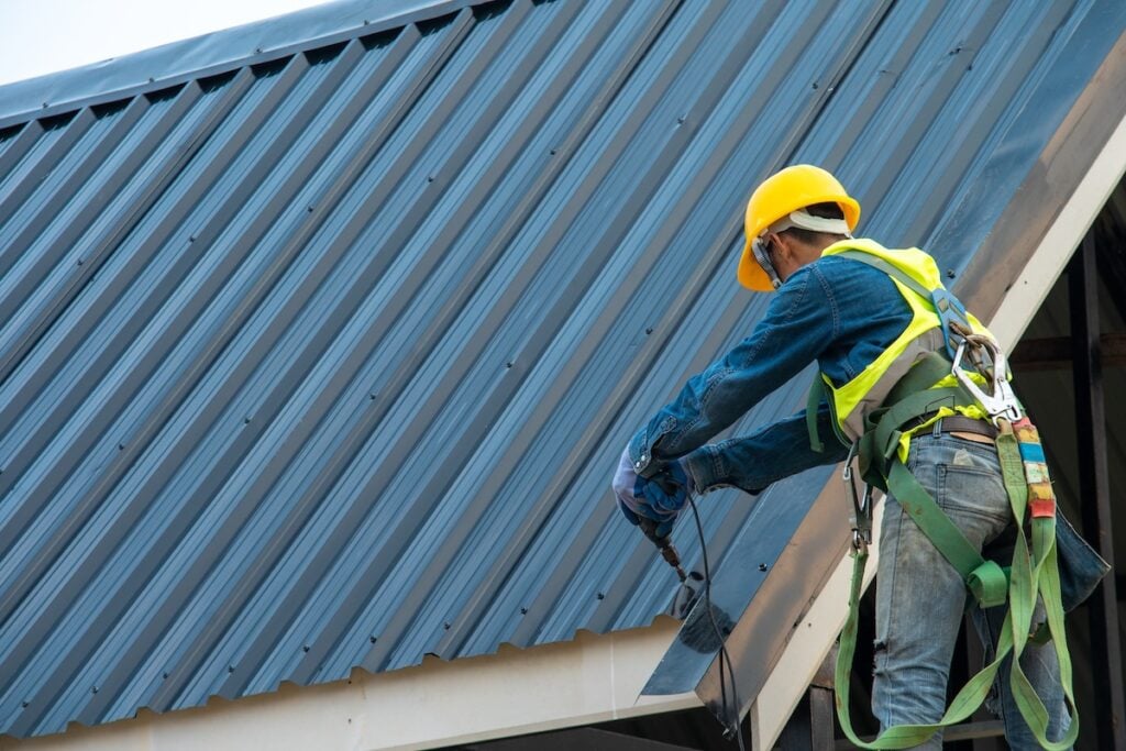 A construction worker in safety gear and a yellow helmet is working on the edge of a metal roof, using tools while wearing a safety harness—demonstrating attention to both safety and the long-term value of durable materials.