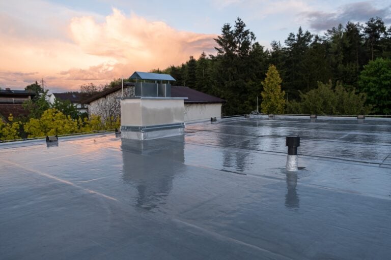 A flat, reflective rooftop with Modified Bitumen Roofing, featuring a chimney and vents, surrounded by trees and houses, under a colorful evening sky with clouds—a scene many homeowners may see in a 2026 Guide to modern roofing solutions.