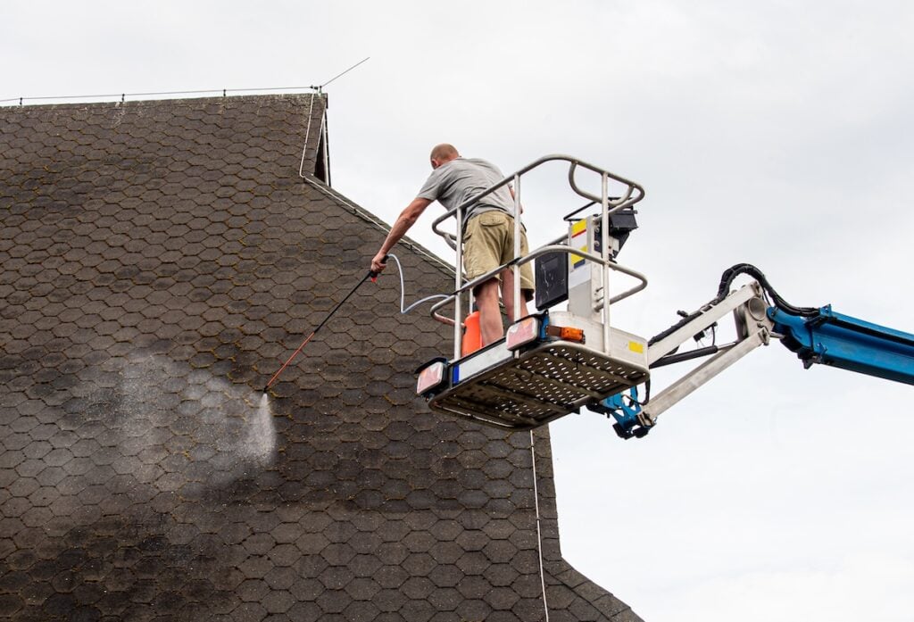 A person standing on a raised platform uses a power washer to remove algae from the shingles on a steep, dark roof against a cloudy sky.