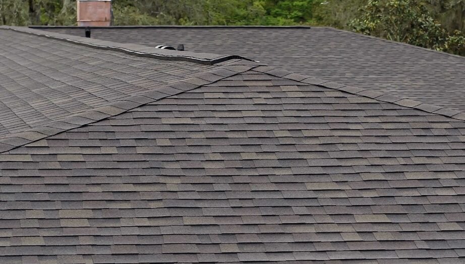 Close-up view of a dark gray shingle roof with overlapping sections, surrounded by trees. Perfect for homeowners curious about 2026 pricing and shingle roof cost, the logo “Shingle Masters Roofing & Construction” appears in the lower right corner.