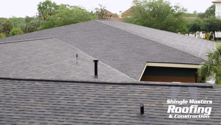 A large, gray shingle roof with vents is seen from above, surrounded by trees. The Shingle Masters Roofing & Construction logo is visible in the lower right corner, making it a great example for homeowners interested in learning about shingle roof cost or exploring the 2026 price breakdown for their next roofing project.