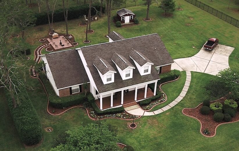 Aerial view of a house with a gray shingle roof, surrounded by green lawn, trees, and garden beds. A curved driveway leads to a parked red car. Logo reads “Shingle Masters Roofing & Construction” in the corner, highlighting quality craftsmanship and cost-effective shingle roof options for your home.