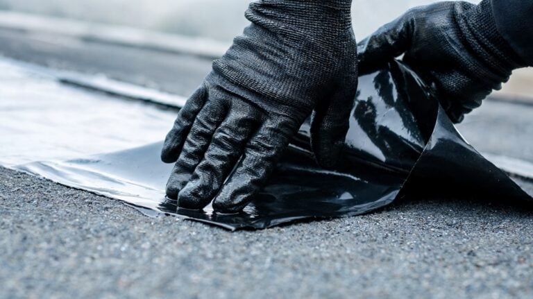 Close-up of construction worker's dark gloved hands peeling and pressing down a glossy black bitumen waterproofing membrane onto a flat asphalt roof surface, showing essential weatherproofing technique.