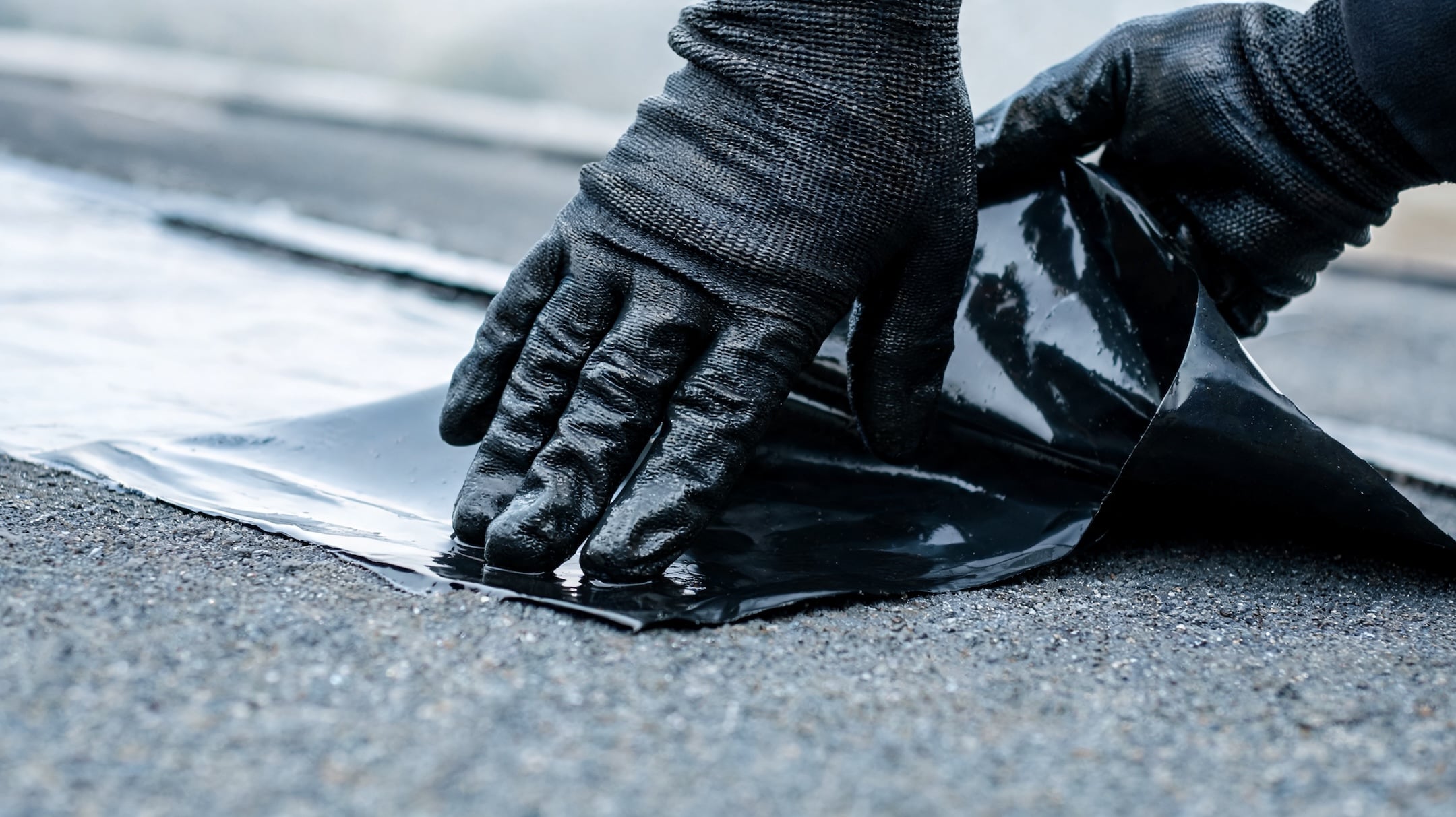 Close-up of construction worker's dark gloved hands peeling and pressing down a glossy black bitumen waterproofing membrane onto a flat asphalt roof surface, showing essential weatherproofing technique.