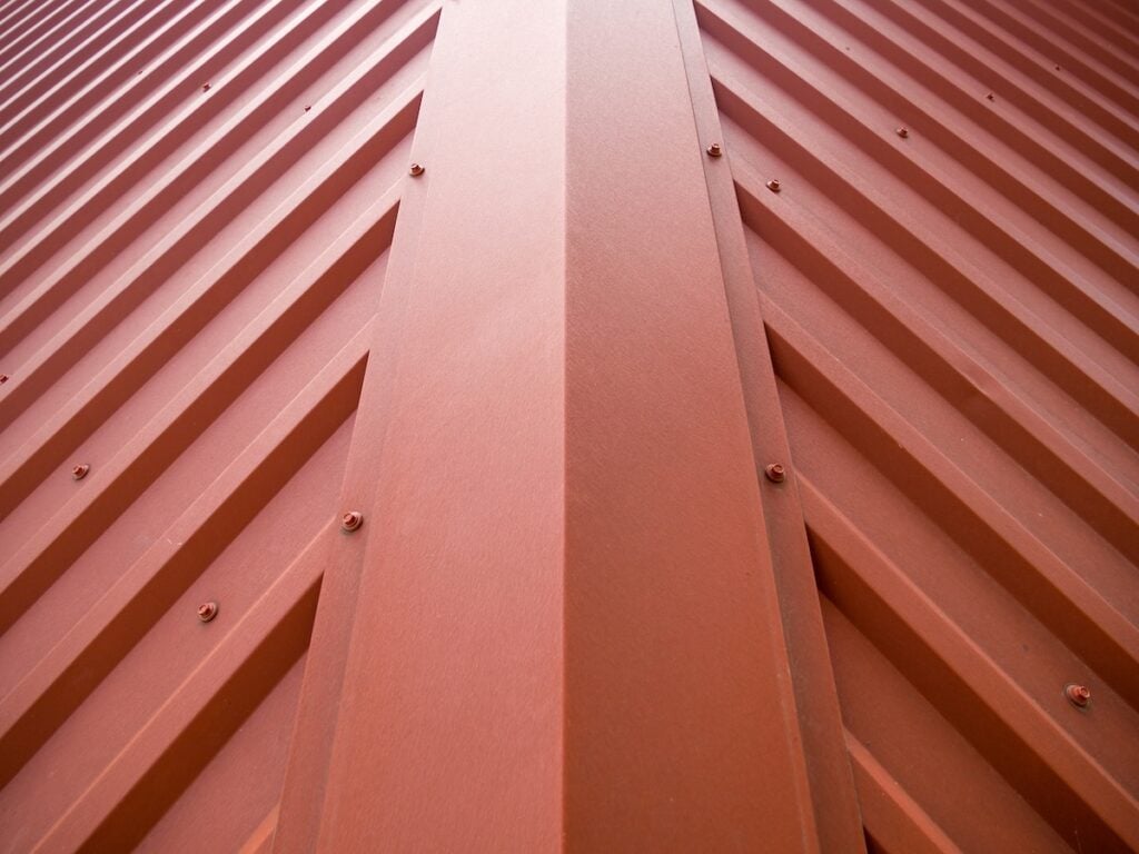 Close-up view of a red metal roof with ridged panels, showing symmetrical lines and screws, creating a geometric pattern converging at the center of the image. The even application of metal roof coating enhances both the durability and vivid color of the panels.