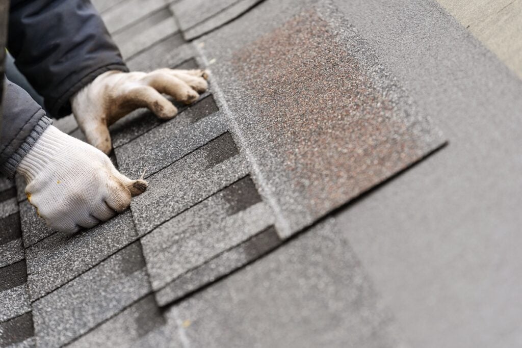 Close-up of gloved hands installing gray asphalt shingles on a roof, aligning them carefully for a secure fit—a detail that many homeowners consider when evaluating shingle roof cost and planning for 2026 pricing.