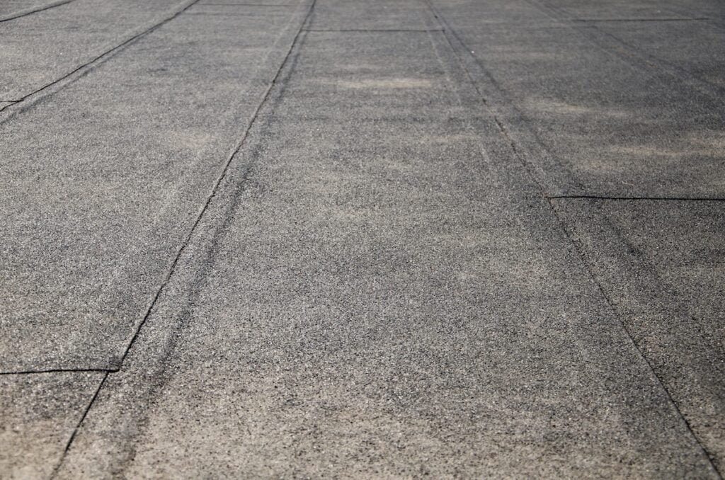 Close-up view of a flat, gray, textured surface with straight black seams—likely a section of a rooftop or pavement—under natural daylight, highlighting potential causes that homeowners might overlook when inspecting for a flat roof leak.