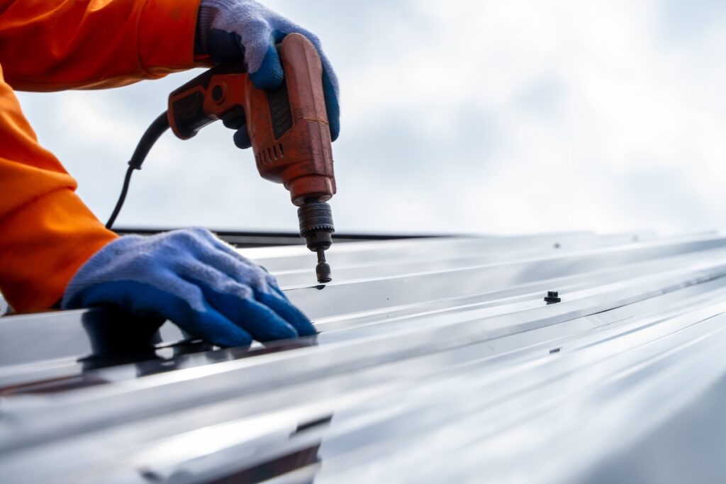 A person wearing blue gloves and an orange jacket uses a power drill to secure a screw into a metal roof sheet outdoors, ensuring durability and value—an important step in managing overall roof cost.