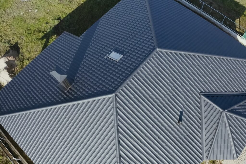 Aerial view of a modern house roof with dark gray metal panels showcasing a stylish metal roof, featuring a skylight, two chimneys, and sections meeting at sharp angles, all surrounded by green grass.