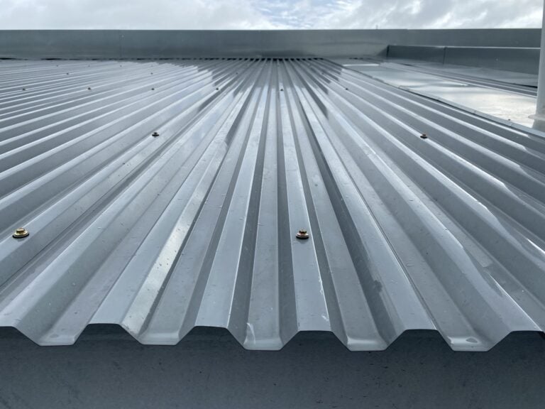 Close-up view of a gray corrugated metal roof with screws securing the panels, under a cloudy sky. The surface appears well-maintained, suggesting the use of a quality metal roof coating for added durability and protection.