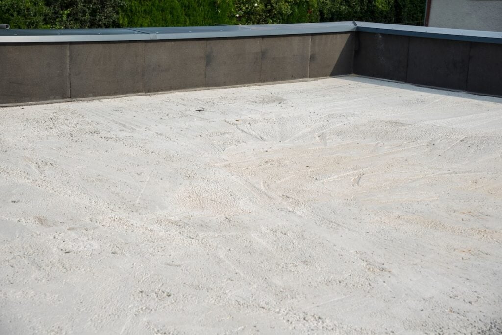 A flat rooftop covered with a layer of light-colored gravel or sand, surrounded by a low, dark parapet wall—common among homeowners—can be vulnerable to the causes of flat roof leaks, even with greenery visible in the background.