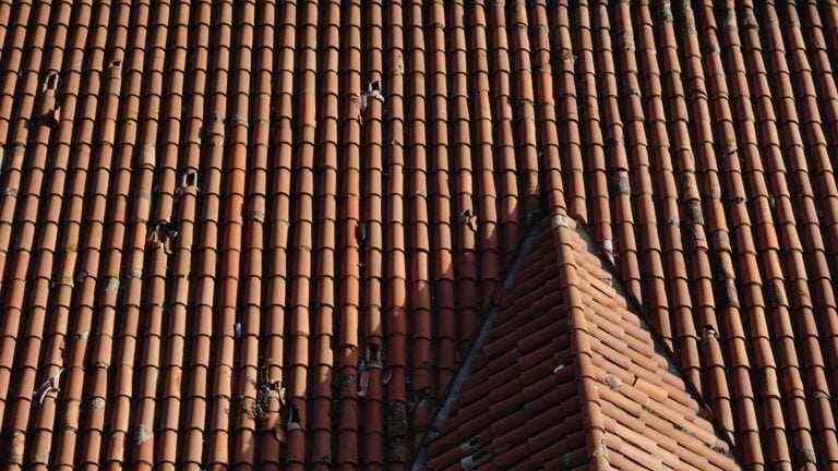 A close-up view of a red clay tiled roof with one triangular section protruding upward. Some tiles show damage and variation in color, creating an uneven and worn appearance—signs that severe weather may have necessitated roof repair or even specialized tile roof storm repair.