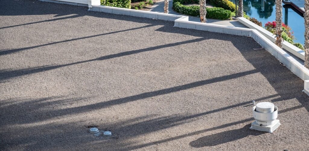 A flat gravel rooftop with a vent and two small fixtures, casting long palm tree shadows—a scene that highlights the importance of flat roof maintenance for preserving both appearance and functionality. Colorful landscaping and water can be seen beyond the rooftop’s edge, offering a glimpse into how proper care can extend your flat roof lifespan.