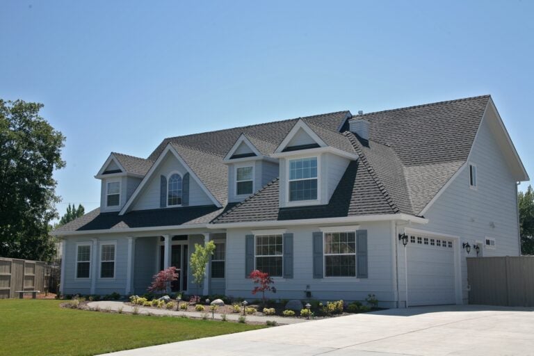 A two-story suburban house with gray siding, white trim, multiple gables, and a durable shingle roof stands under a clear blue sky. Homeowners will appreciate the well-maintained front yard and the convenience of an attached two-car garage, as well as having insight into 2026 pricing for shingle roof cost when planning future upgrades.