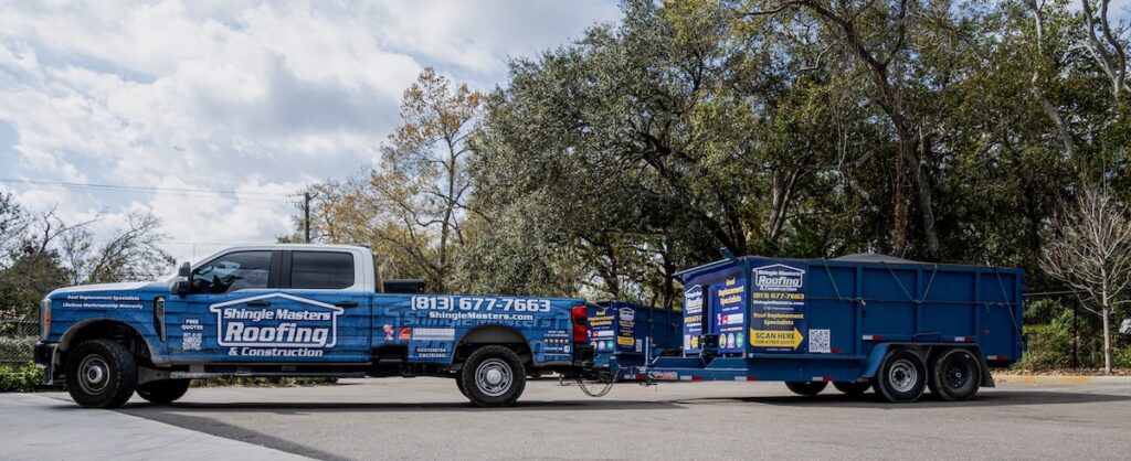 A Shingle Masters Roofing & Construction truck, wrapped in company branding, is parked and hitched to a large blue trailer with signs promoting 3-Tab and Architectural Shingles. Trees and a cloudy sky are visible in the background, highlighting the versatile roofing solutions offered by the company.