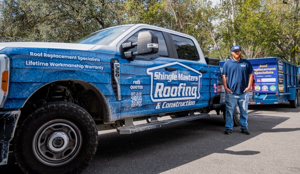 A man stands next to a blue Shingle Masters Roofing & Construction truck, which advertises roof replacement specialist services. The matching trailer parked behind them highlights their expertise in flat roof maintenance and roof maintenance tips. Trees are visible in the background, creating a professional and approachable atmosphere.