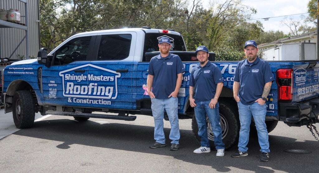 Three men in blue shirts and hats stand next to a blue Shingle Masters Roofing & Construction truck, parked outside on a sunny day. Specializing in services like metal roof coating, the team is ready to handle your roofing needs. Trees and a building are visible in the background.