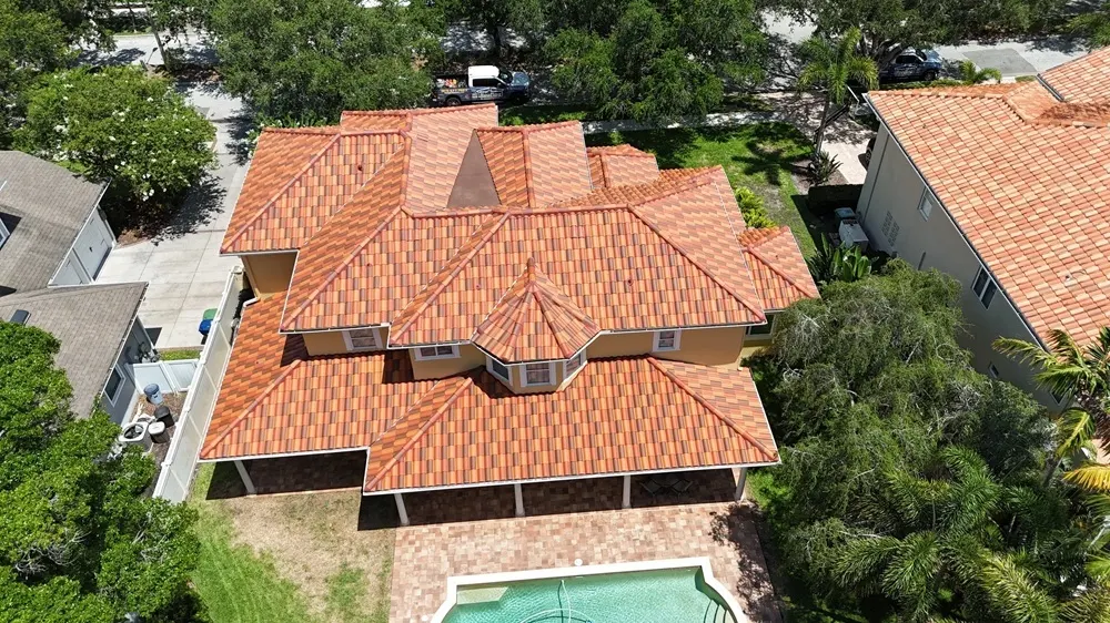 Aerial view of a large house with a patterned red tile roof, surrounded by trees and neighboring homes, with a swimming pool in the backyard—a beautiful property that may benefit from timely tile roof repair to prevent issues caused by severe weather roof damage.