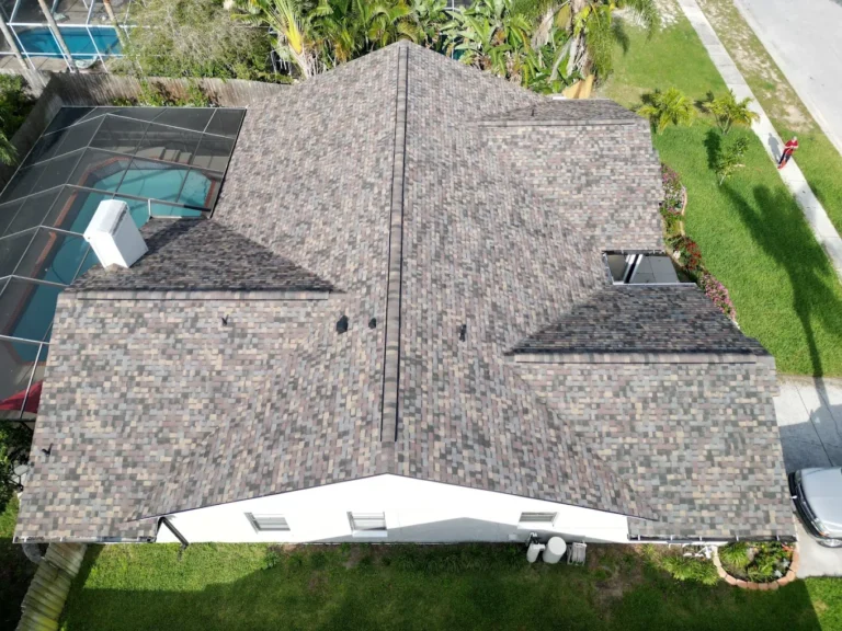 Aerial view of a house with a newly shingled roof featuring durable architectural shingles in gray and brown tones, surrounded by green grass, trees, and a screened backyard pool area on the left side.