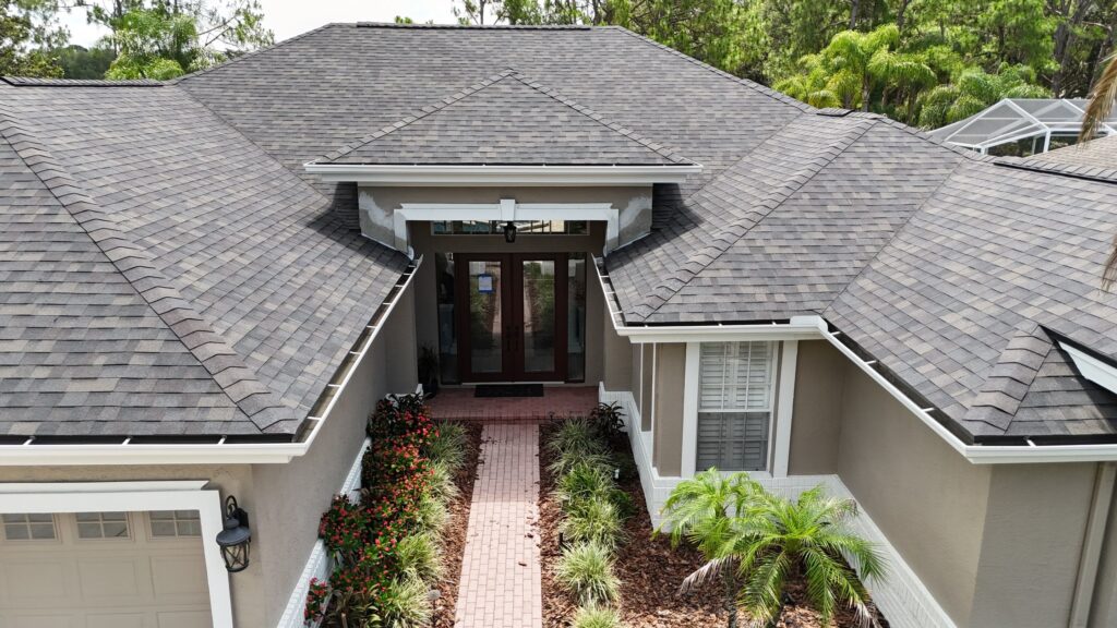 impact resistant shingles Aerial view of a single-story house with gray impact resistant shingles, beige walls, white trim, a brick walkway, landscaped plants, and large glass double doors at the entry—showcasing features that are worth any extra cost for enhanced durability and style.