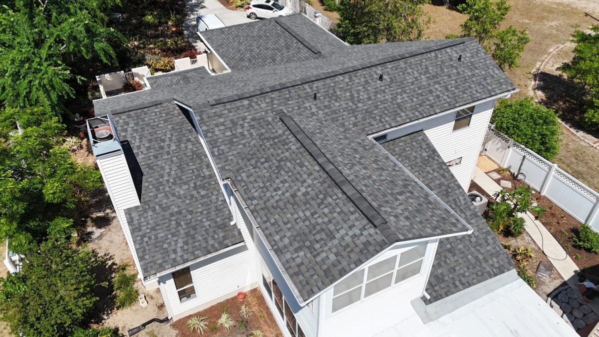 impact resistant shingles Aerial view of a house with a dark gray Impact Resistant Shingles roof, surrounded by trees and landscaping. The roof has multiple ridges and sections, forming an irregular shape, showcasing that the extra cost is often worth it for added durability and protection.