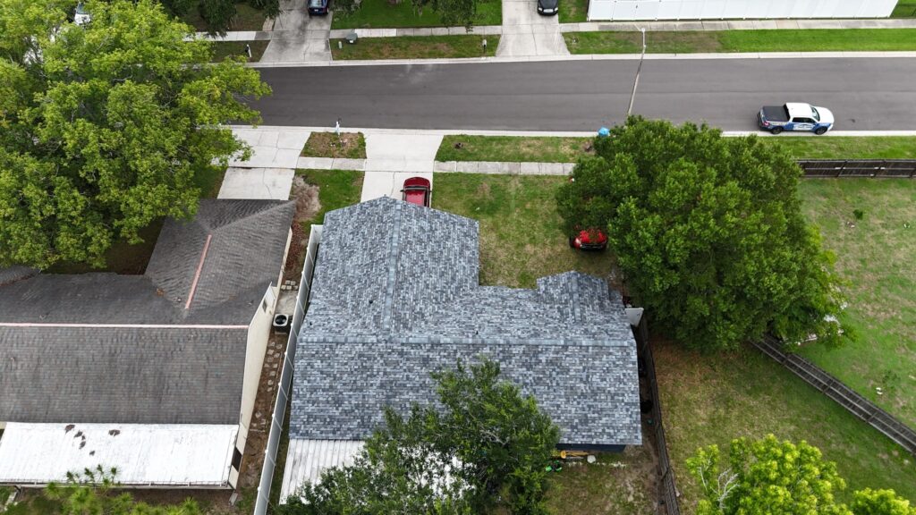 impact resistant shingles Aerial view of a gray-roofed house featuring impact resistant shingles, with trees on both sides, a red car in the driveway, and a street with a parked white vehicle in a suburban neighborhood. While these roofing materials may involve extra cost, they offer added durability and protection for your home.