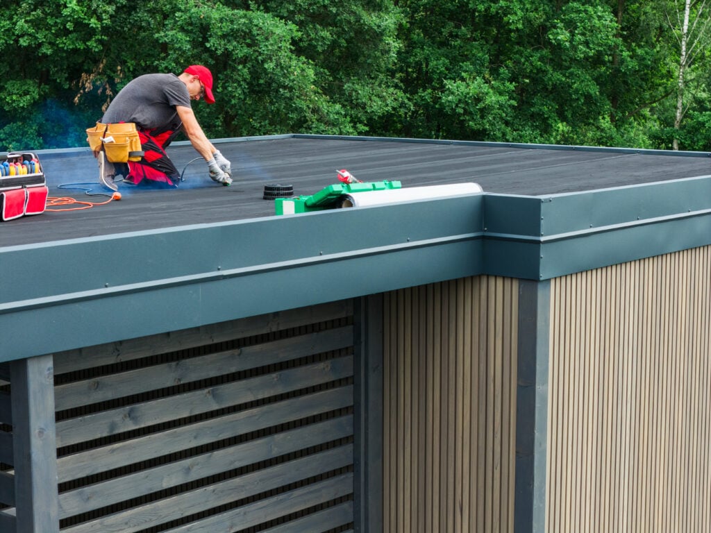 flat roof pitch A worker in a red cap and gloves installs or repairs a flat roof with a slight flat roof pitch on a modern building surrounded by trees, using tools and materials placed nearby.