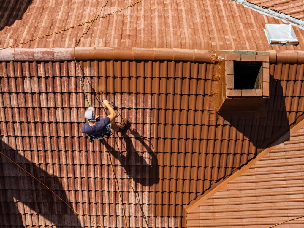 A worker wearing a helmet and harness is cleaning or repairing a terracotta-tiled roof, using safety ropes for protection. There is a rectangular chimney and a white hatch visible on the roof. This maintenance work is essential to prepare the surface for tile roof coating, which helps extend the lifespan of the tiles and enhances weather resistance—just some of the benefits of tile roof coating that homeowners can enjoy.