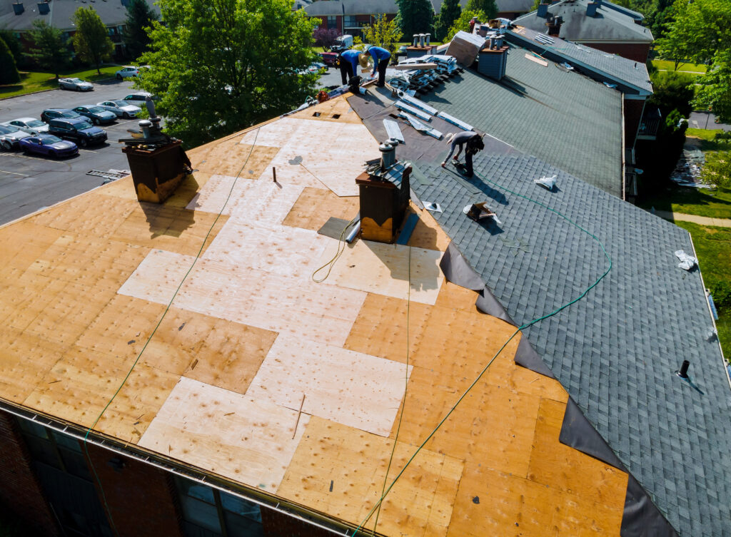 Workers are repairing a large building roof, replacing old shingles with new ones to ensure improved shingle roof coverage. Part of the roof has exposed plywood, while other sections have fresh shingles installed. Equipment and materials are scattered around, all contributing to a quality result that may be eligible for a comprehensive roofing warranty.