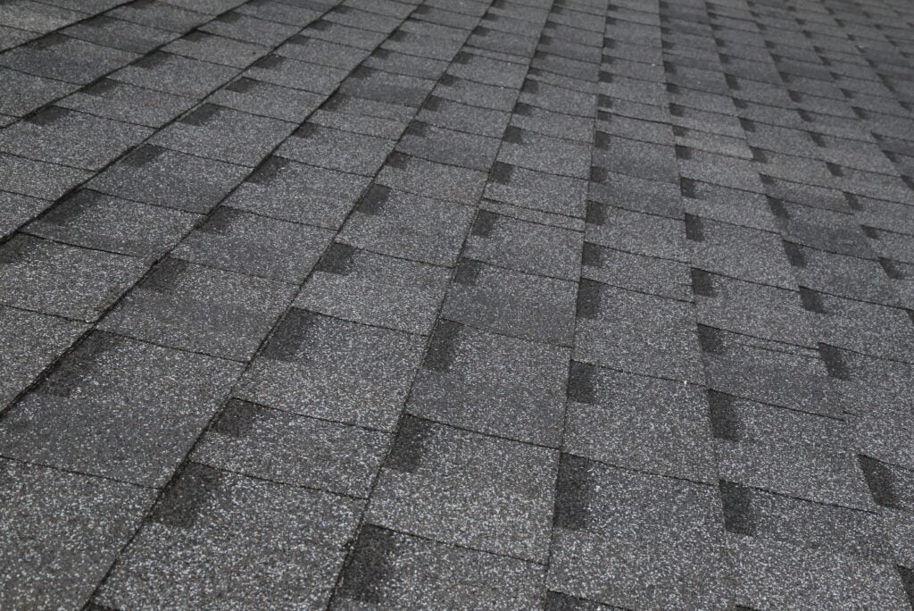A close-up view of gray asphalt shingle roof shingles arranged in a neat, overlapping pattern, creating a textured surface.