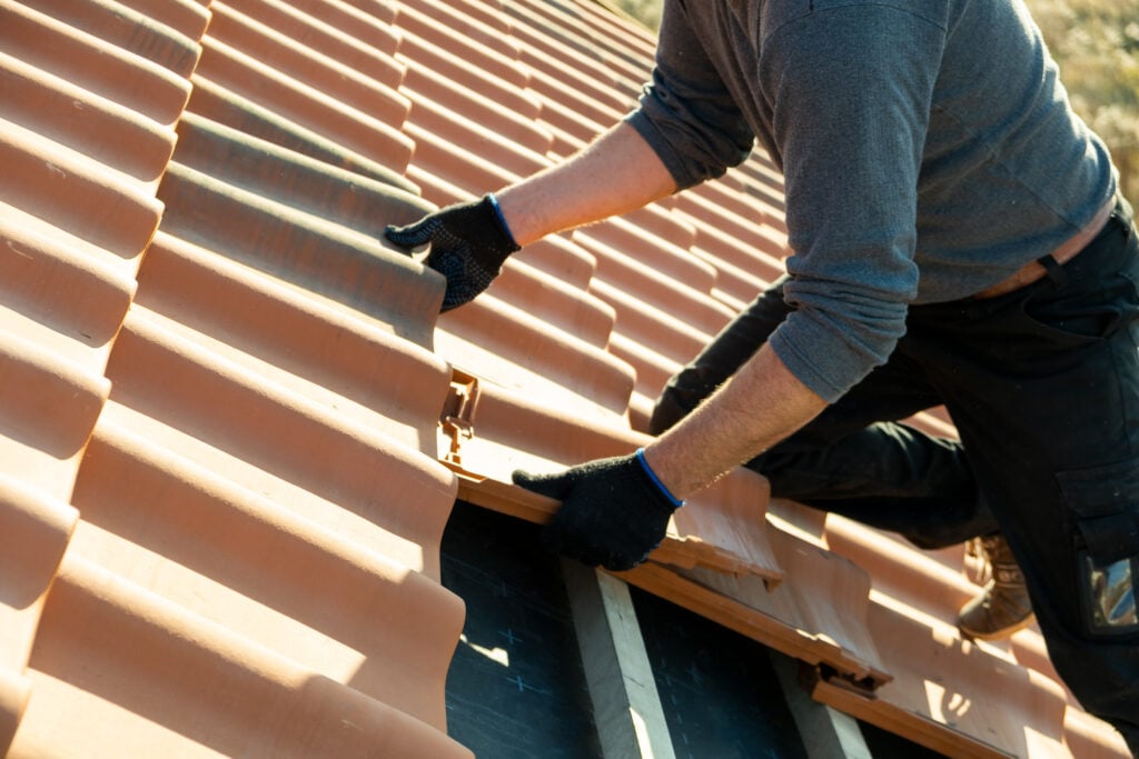 A person wearing gloves installs terracotta roof tiles while kneeling on a partially completed roof, preparing the surface for a protective tile roof coating to enhance durability and weather resistance.