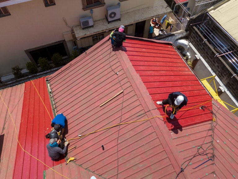 signs you need a new roof Four people work on a red rooftop, handling ropes and tools—perhaps addressing some signs to replace roof materials. One person sits near the peak, while three others are spaced along the roof. Buildings and equipment are visible around and below the rooftop, suggesting an active roof replacement project is underway.