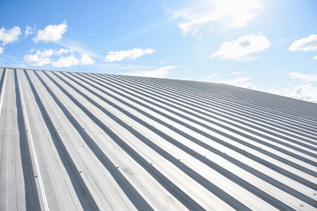 A sleek aluminum roof with parallel ridges is shown under a bright blue sky, scattered clouds drifting above as sunlight reflects brilliantly off the surface.