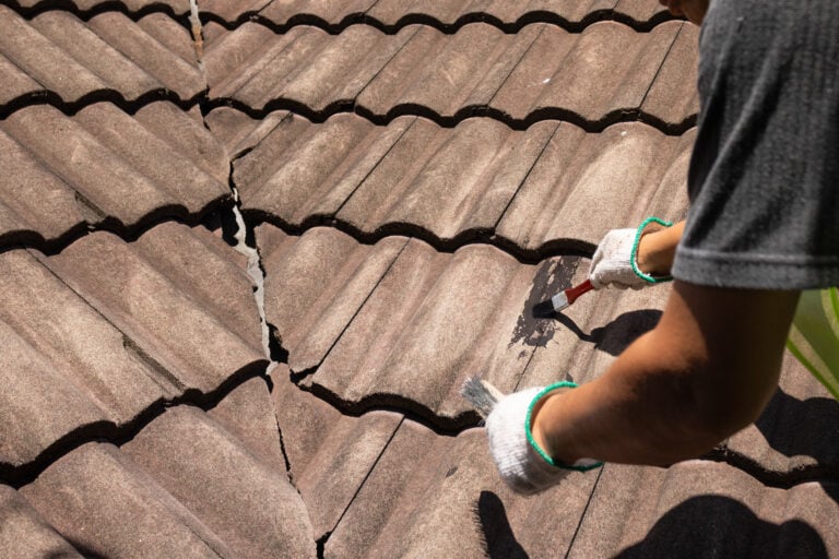 tile roof coating A person wearing gloves repairs a large crack in a tiled roof, using a brush and tool to apply a protective substance as part of routine tile roof maintenance.