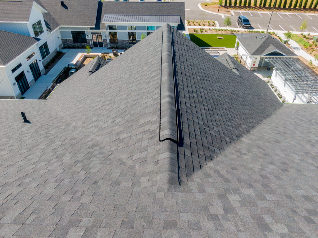 impact resistant shingles Aerial view of a gray shingled roof with a vent ridge, surrounded by modern buildings, green lawns, and a parking lot in the background. The roof features impact resistant shingles, offering added protection that many homeowners find worth the extra cost.