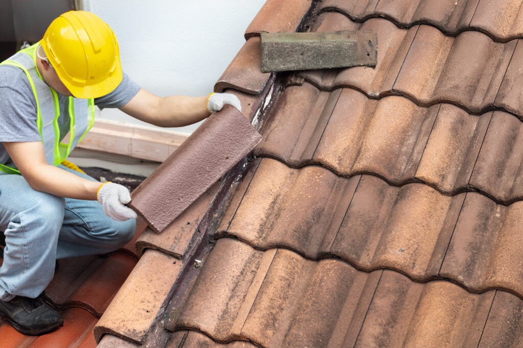 tile roof coating A construction worker wearing a yellow helmet and safety vest repairs a brown tiled roof, lifting one tile to inspect or replace it, demonstrating the importance of regular tile roof maintenance.
