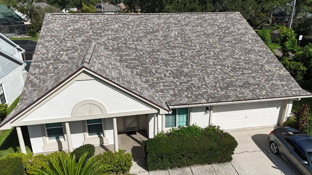 Aerial view of a house with a shingle roof, beige exterior, two-car garage, and driveway with a car parked. Bushes line the walkway to the front door, highlighting the curb appeal and durability that comes with reliable shingle roof warranty coverage.
