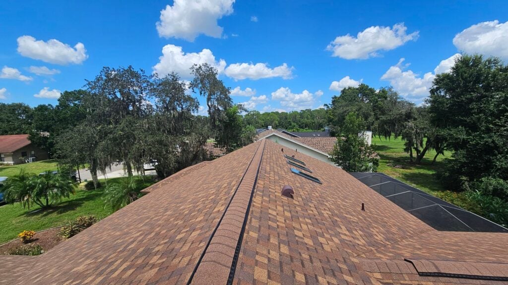 View from the peak of a brown shingle roof with trees, lawns, and several houses under a bright blue sky filled with fluffy clouds—a picturesque scene that could inspire any 2026 guide to Florida homes.