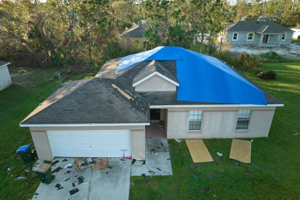 signs you need a new roof A house with a blue tarp covering part of its roof, likely due to storm damage—a definite roof sign that you may need to replace your roof—debris scattered on the lawn and driveway, and neighboring houses visible in the background.