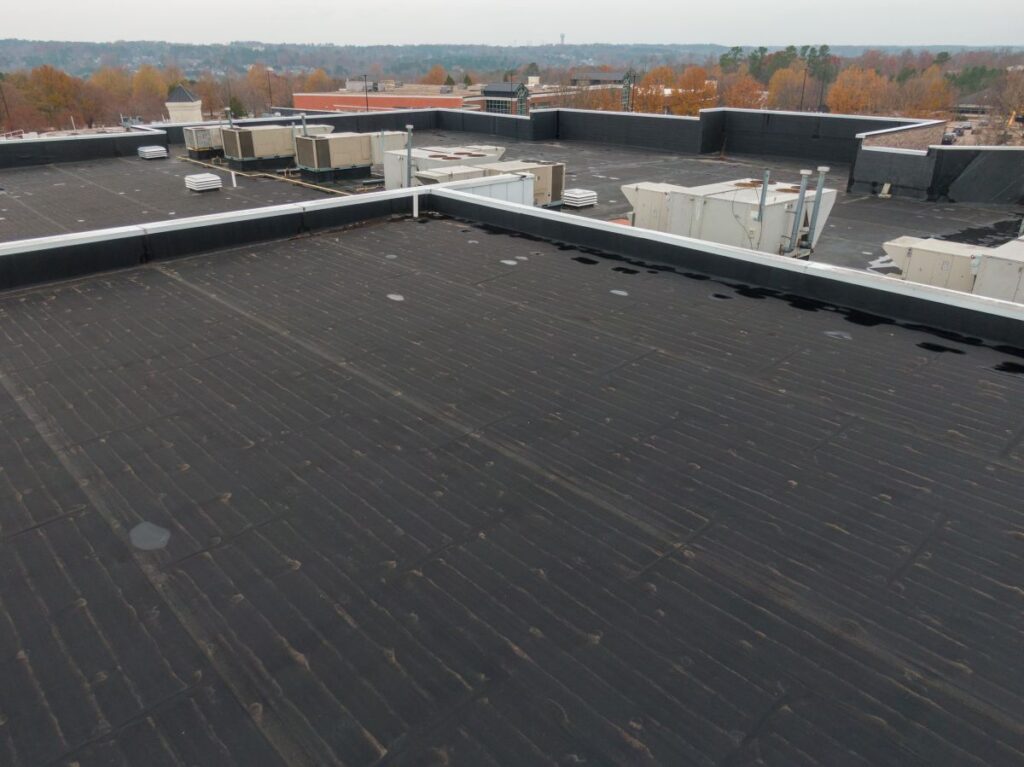 Flat commercial rooftop with black waterproof membrane, several HVAC units, and low surrounding walls. Unlike a shingle roof or metal roof, this flat surface ensures durability in various weather conditions. Trees with autumn foliage and distant buildings are visible in the background under a cloudy sky.