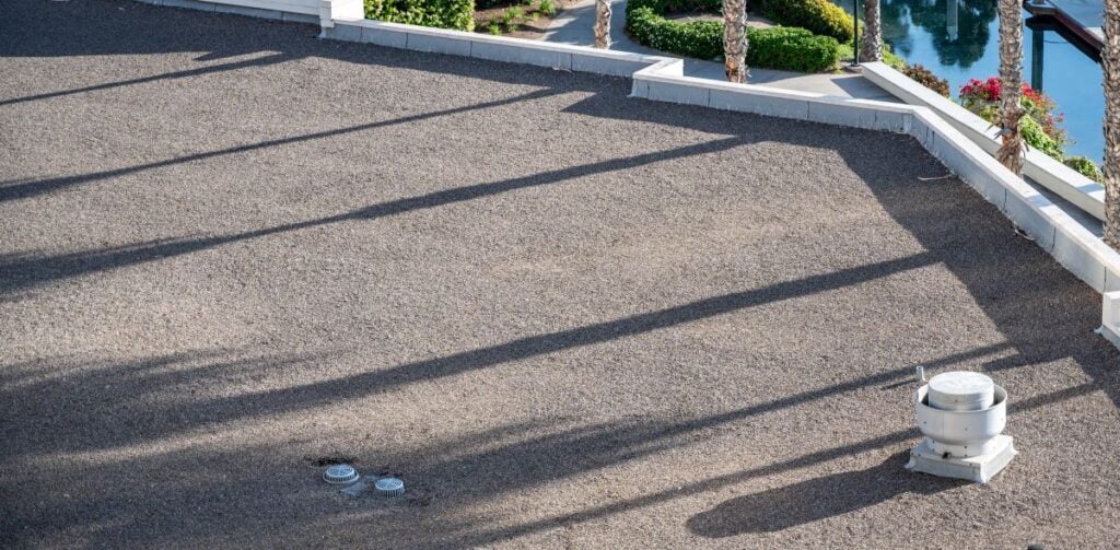 flat roof pitch A flat gravel rooftop with a gentle flat roof slope features a vent and two small capped pipes. Shadows of tall palm trees stretch across the rooftop. In the background, there are landscaped gardens and water.