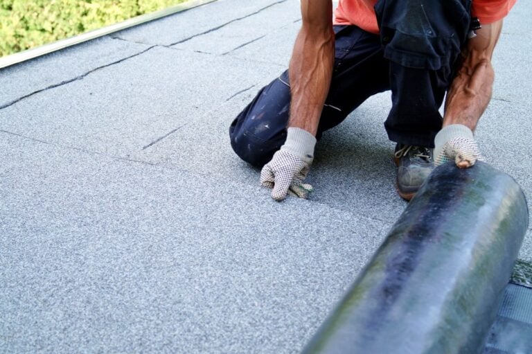 flat roof pitch A person wearing work gloves kneels on a flat roof while unrolling a sheet of roofing material during construction or repairs. Only their arms and part of their body are visible, emphasizing the hands-on nature of working on a rooftop with minimal roof pitch.