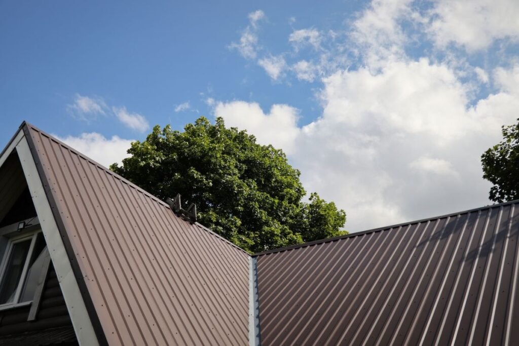 metal roof vs shingle roof hurricane A brown metal roof with a triangular peak is shown against a background of green trees and a partly cloudy blue sky, offering durable protection that stands strong even during hurricane conditions.