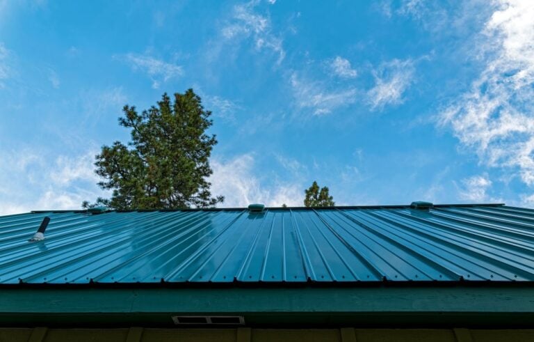 metal roof vs shingle roof hurricane A green metal roof of a building is shown with trees and a blue sky with wispy clouds in the background, highlighting the durability of a metal roof even in hurricane-prone areas.