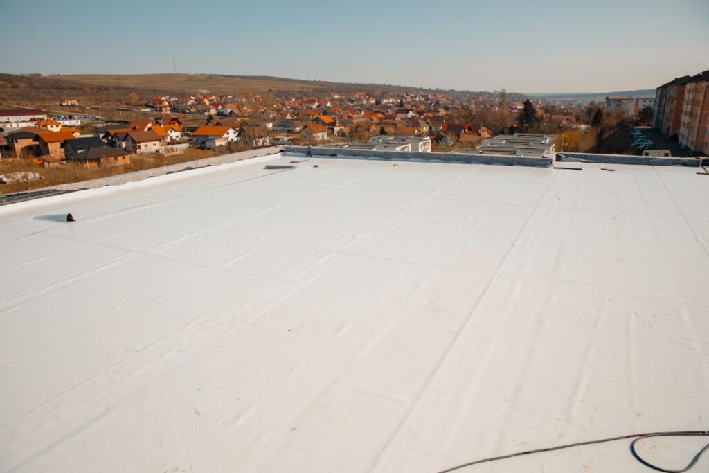 best flat roof material A large white flat rooftop with a smooth surface overlooks a residential area with shingle roofs, fields, and distant hills under a clear sky.