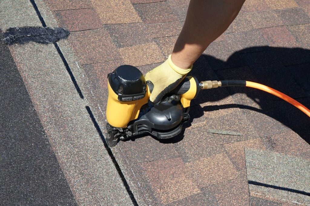 signs you need a new roof A person wearing a yellow glove uses a nail gun to secure brown asphalt shingles on a roof, illustrating the careful process involved in a roof replacement. An orange air hose is attached to the tool.