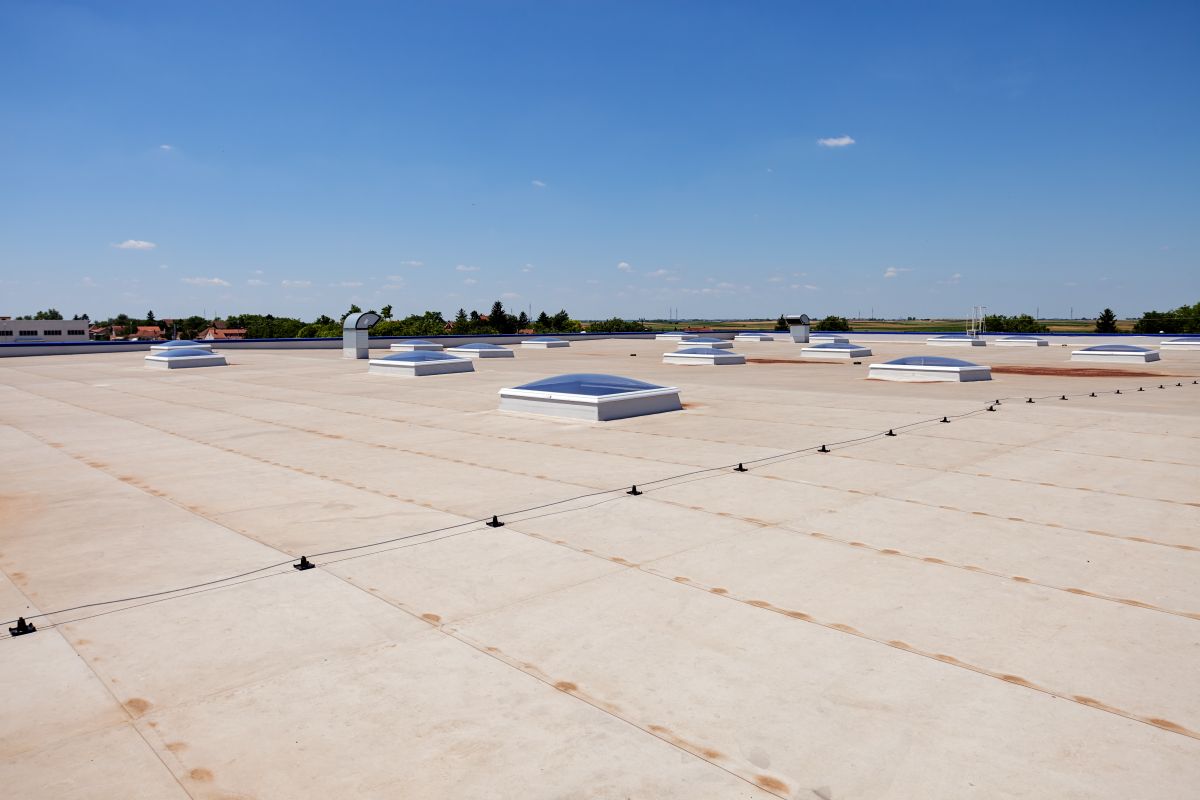 best flat roof material Flat commercial building rooftop with multiple skylights and ventilation units on a sturdy metal roof, under a clear blue sky. Trees and other buildings are visible in the background.